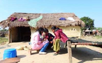A female journalist speaks with two young women while seated outside a home with a thatched roof in Lumbini Province, Nepal.