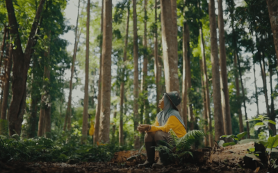 A woman wearing a yellow shirt, boots, and a traditional headscarf and hat sits on the forest ground, overlooking trees.