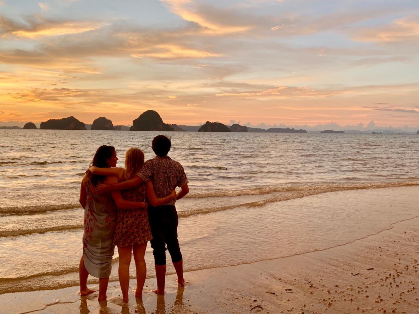 Three people on a beach enjoy the sunset with their arms around one another