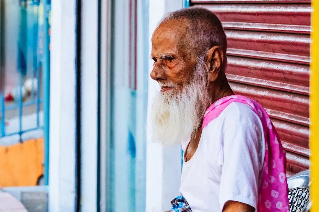 A bearded elderly man sits by a garage door in Sri Lanka.