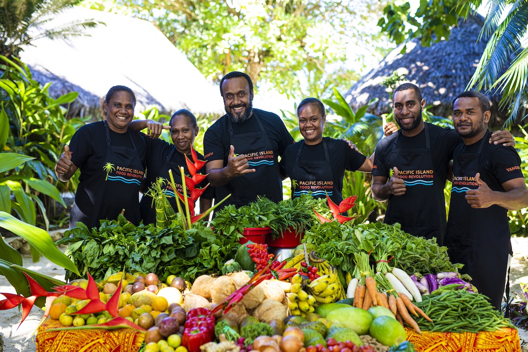 Five participants of the Pacific Island Food Revolution stand behind a cornucopia of Pacific Island vegetables and grains.