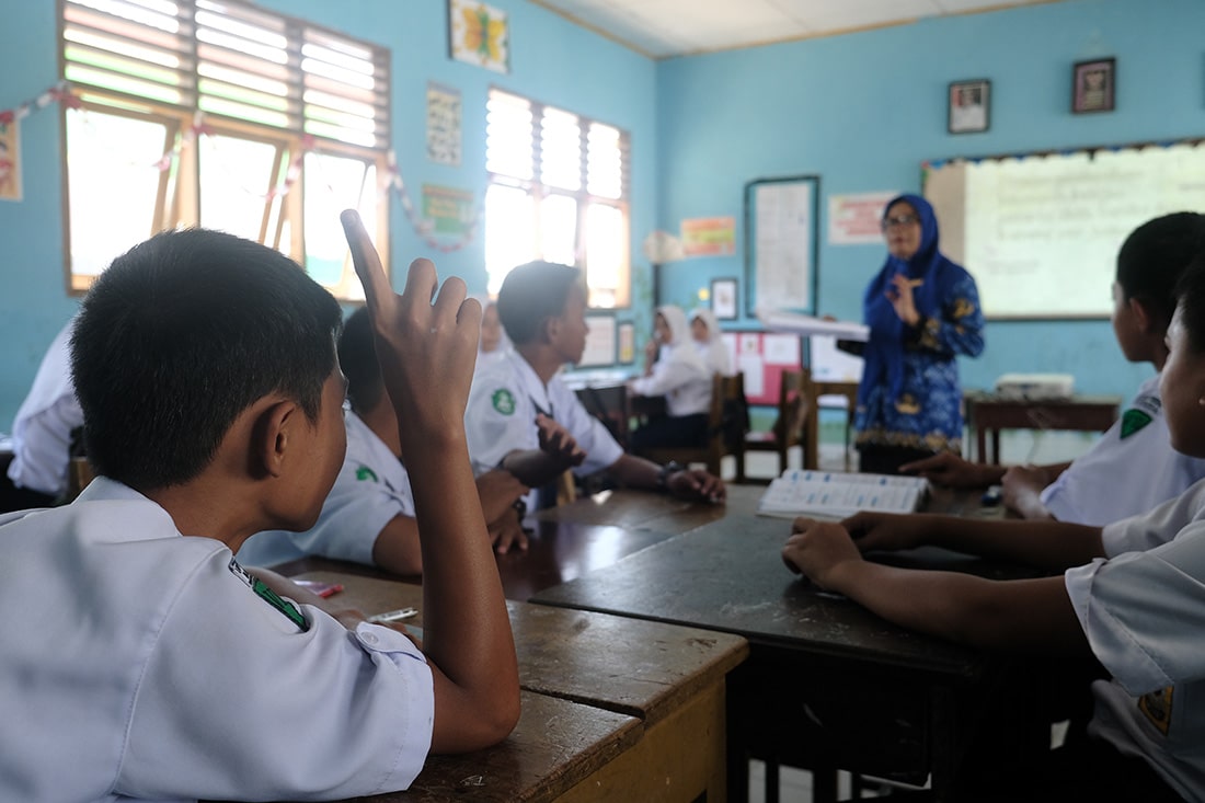 A boy in a school uniform raises his hand in a classroom.