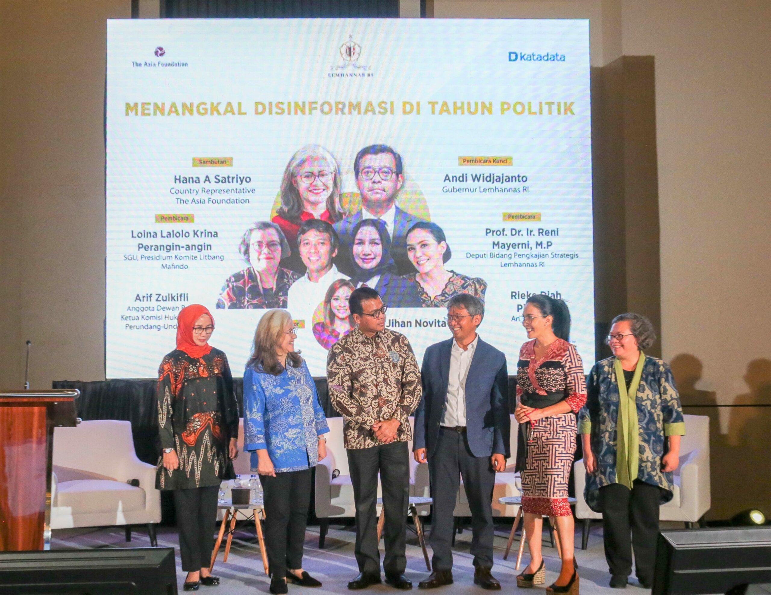 A group of men and women at an event stand on stage in front of large screen with a promotional graphic