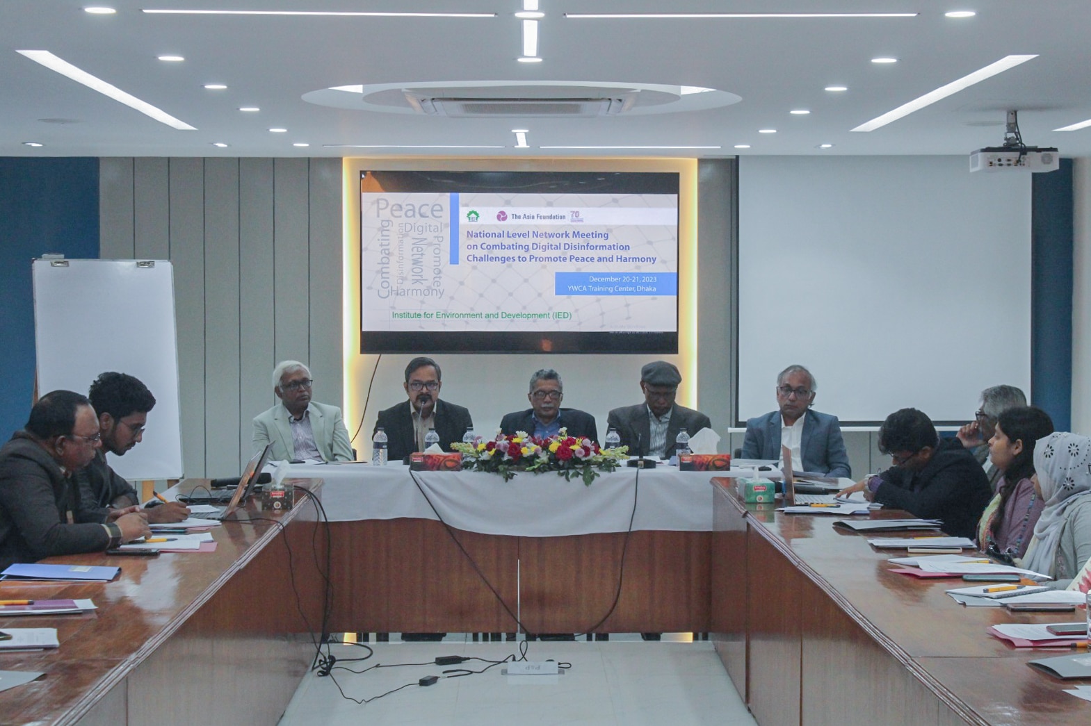 Members of various Bangladeshi organizations sit at one side open rectangular table. The camera faces the members and a screen noting the meetings formal title.
