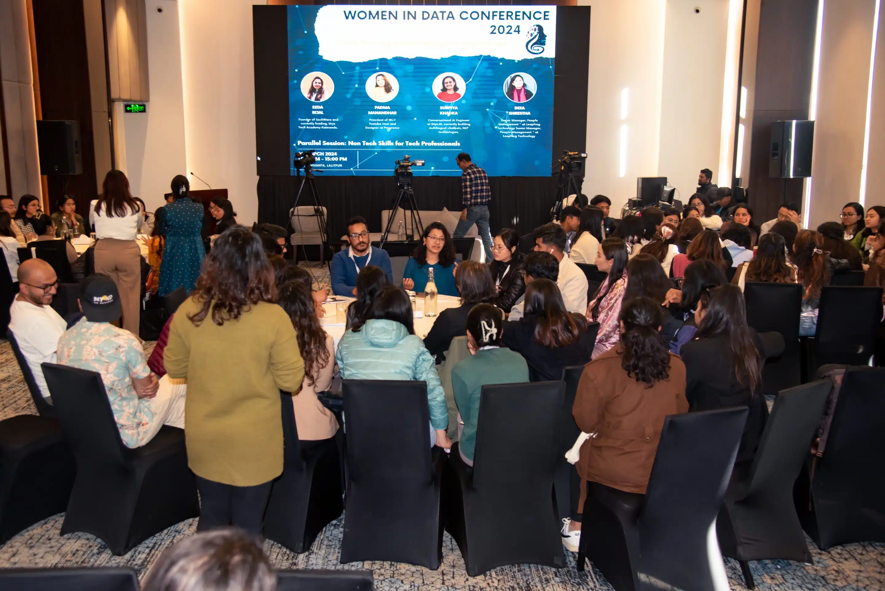A large group sit in a circle at a roundtable. There is a Women in Data Conference panel session graphic on the screen behind them.